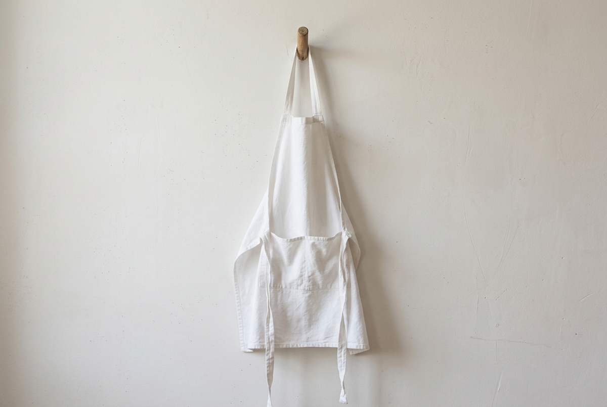 Tablier blanc en coton épais suspendu à une patère en bois contre un mur clair